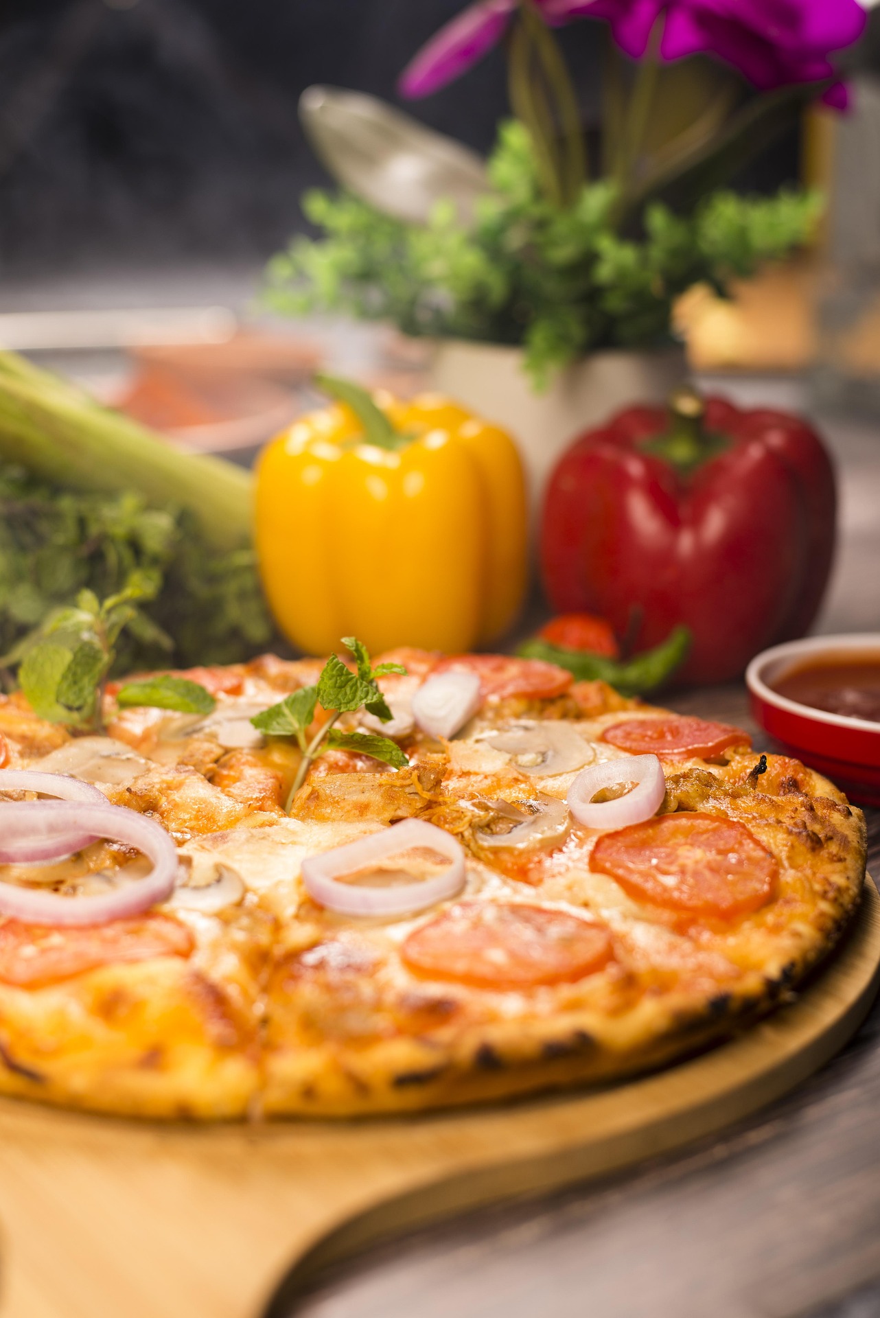 Pizza op pizzaplank met potje saus en paprika's en diverse groenten er rond.
